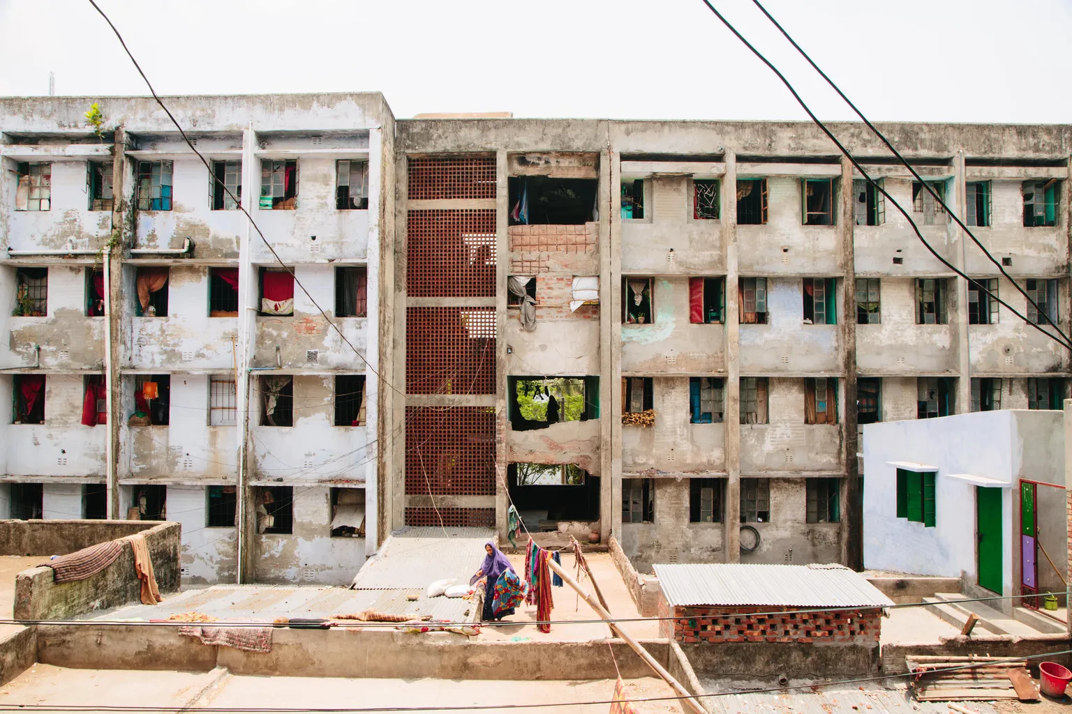 A weathered multi-story apartment building with open windows and laundry hanging, viewed from a nearby rooftop with a person sitting.