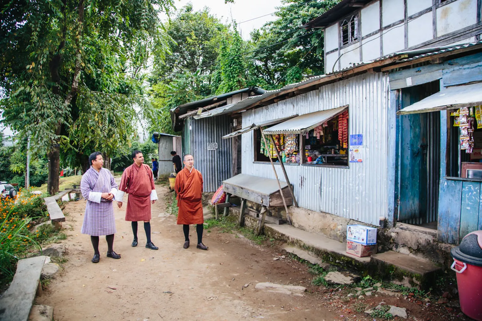 Three men in traditional attire stand outside a small shop with a corrugated metal exterior, surrounded by greenery and a dirt path.