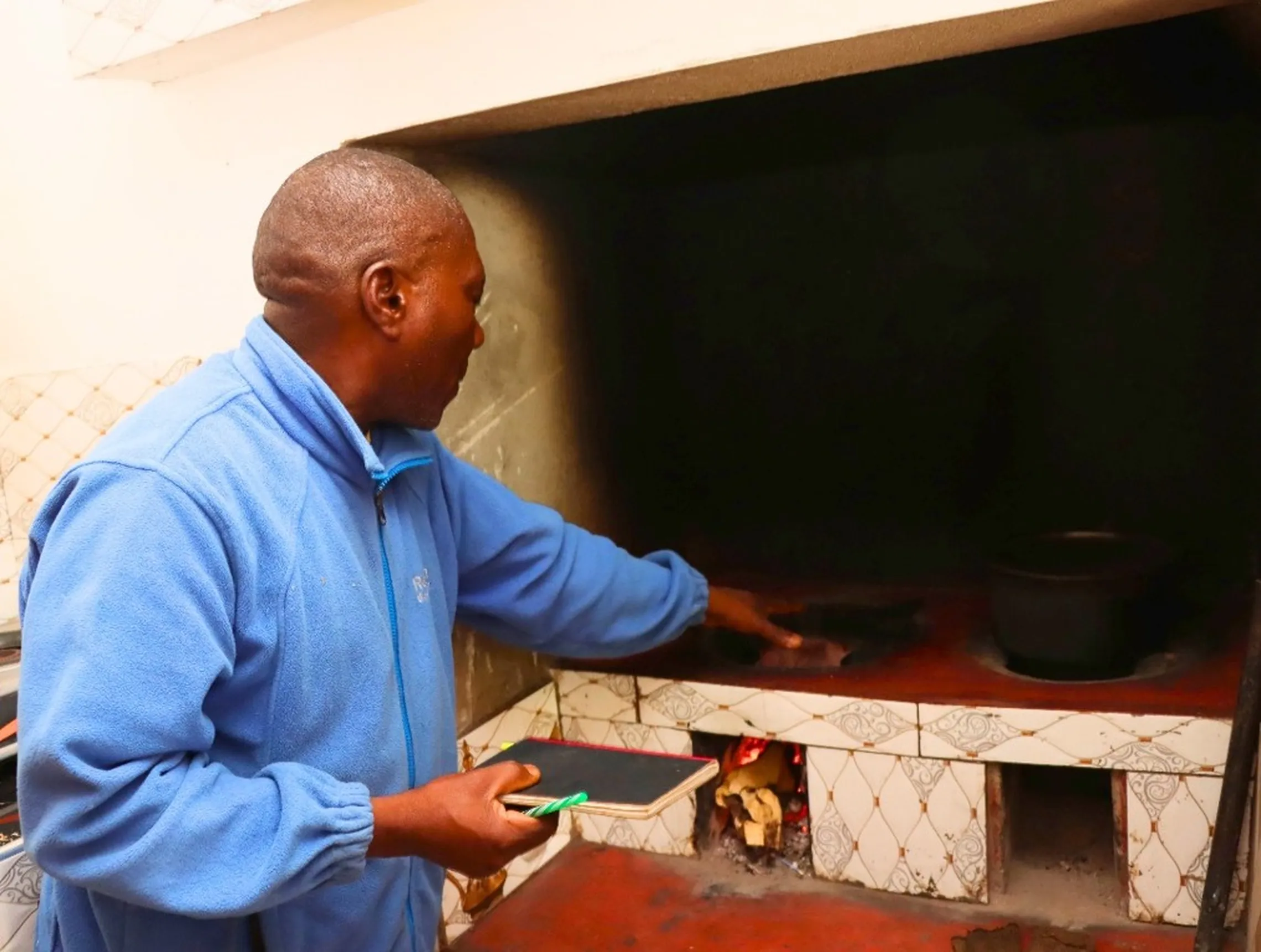 A person in a blue jacket tends to a wood-fired stove, with a pot on top and flames visible inside the stove.