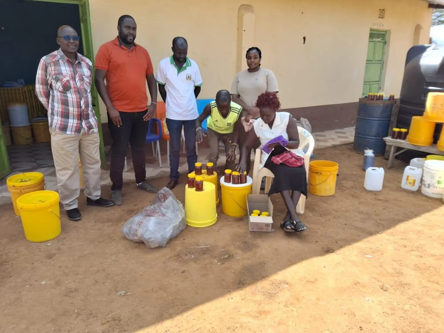 Six people organize honey bottles outside a building, surrounded by yellow buckets and containers on the ground.