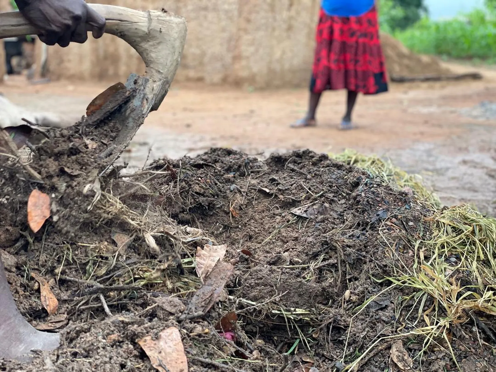 Close-up of a person using a hoe to turn over soil and organic matter, with another person standing in the background on a dirt path.