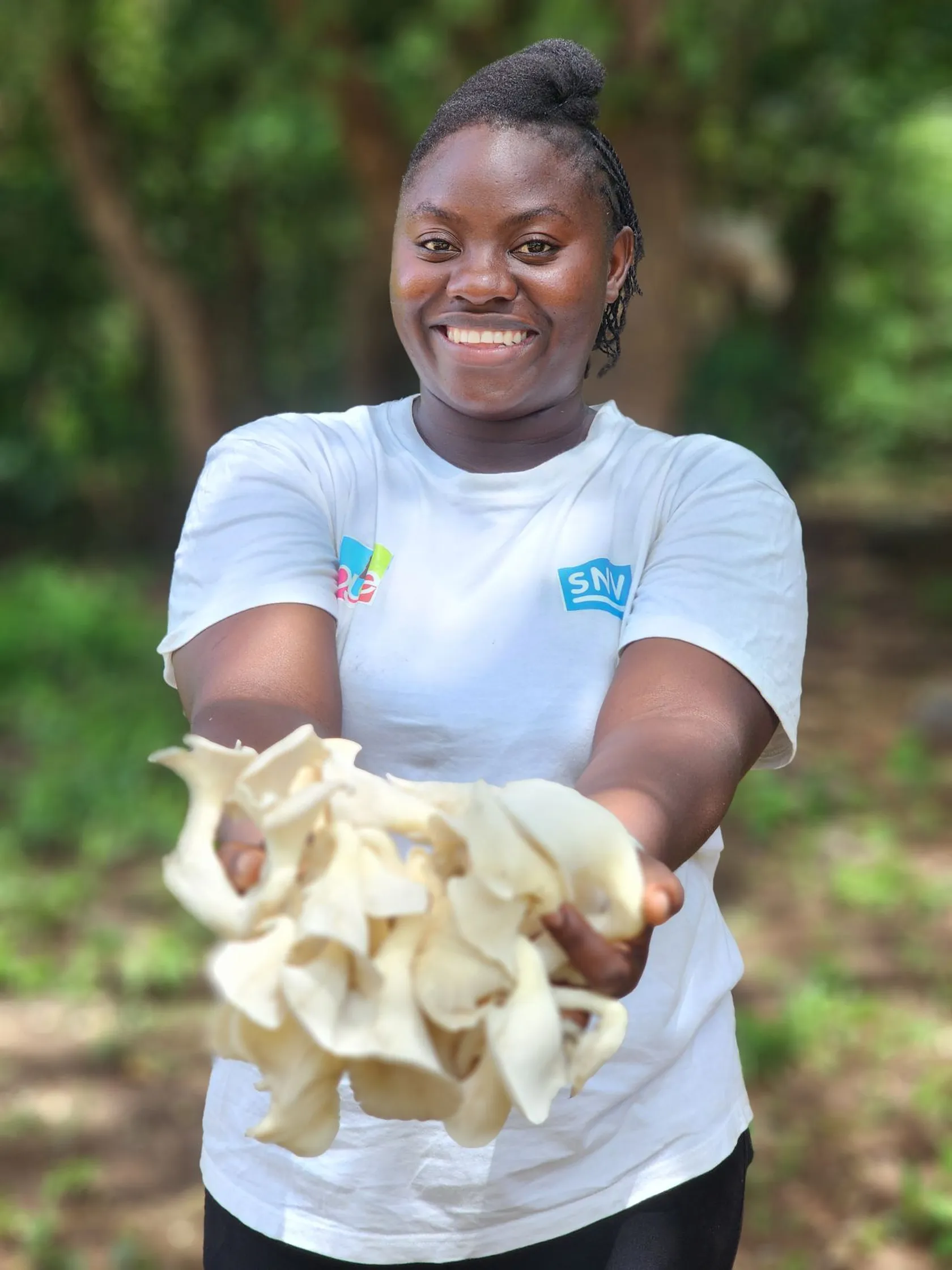 Smiling woman in a white shirt holds a bunch of fresh mushrooms outwards in a green, outdoor setting.
