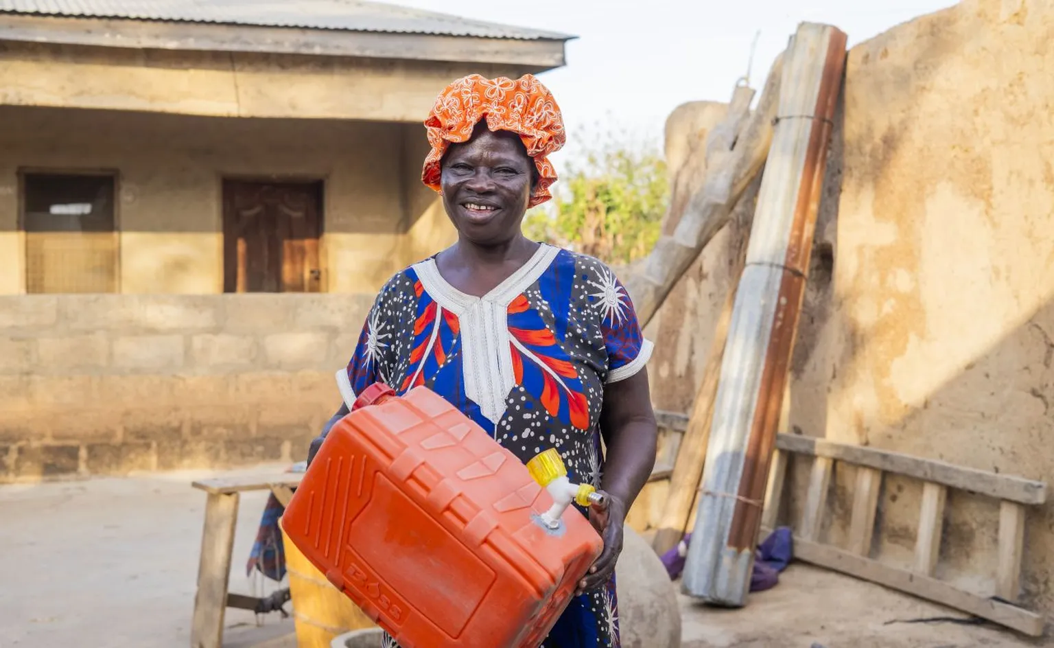 Woman in colorful dress and orange headscarf smiling, holding a large red container outdoors near a building with a metal roof.