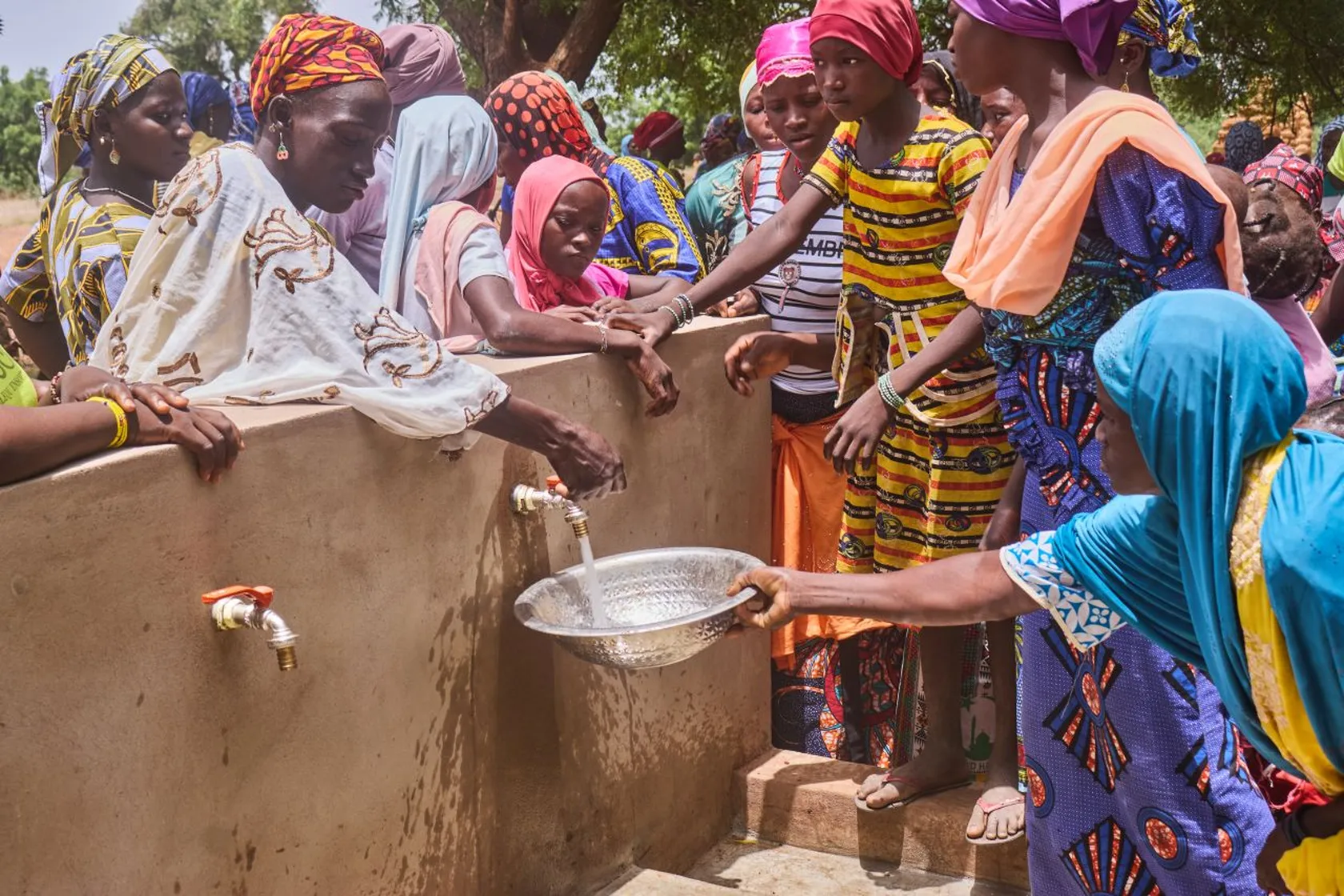 A group of women and children gather around a public water faucet, filling a bowl, in a rural outdoor setting.