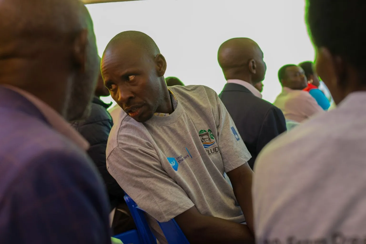 A man in a light gray T-shirt with a logo listens to another person in a seated group setting, with more people in the background.