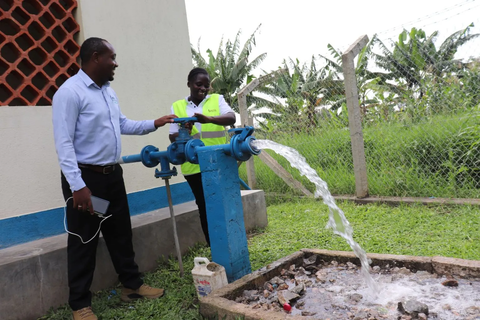 Two people operate a water pump, releasing a strong flow of water into a concrete basin outdoors, with greenery in the background.