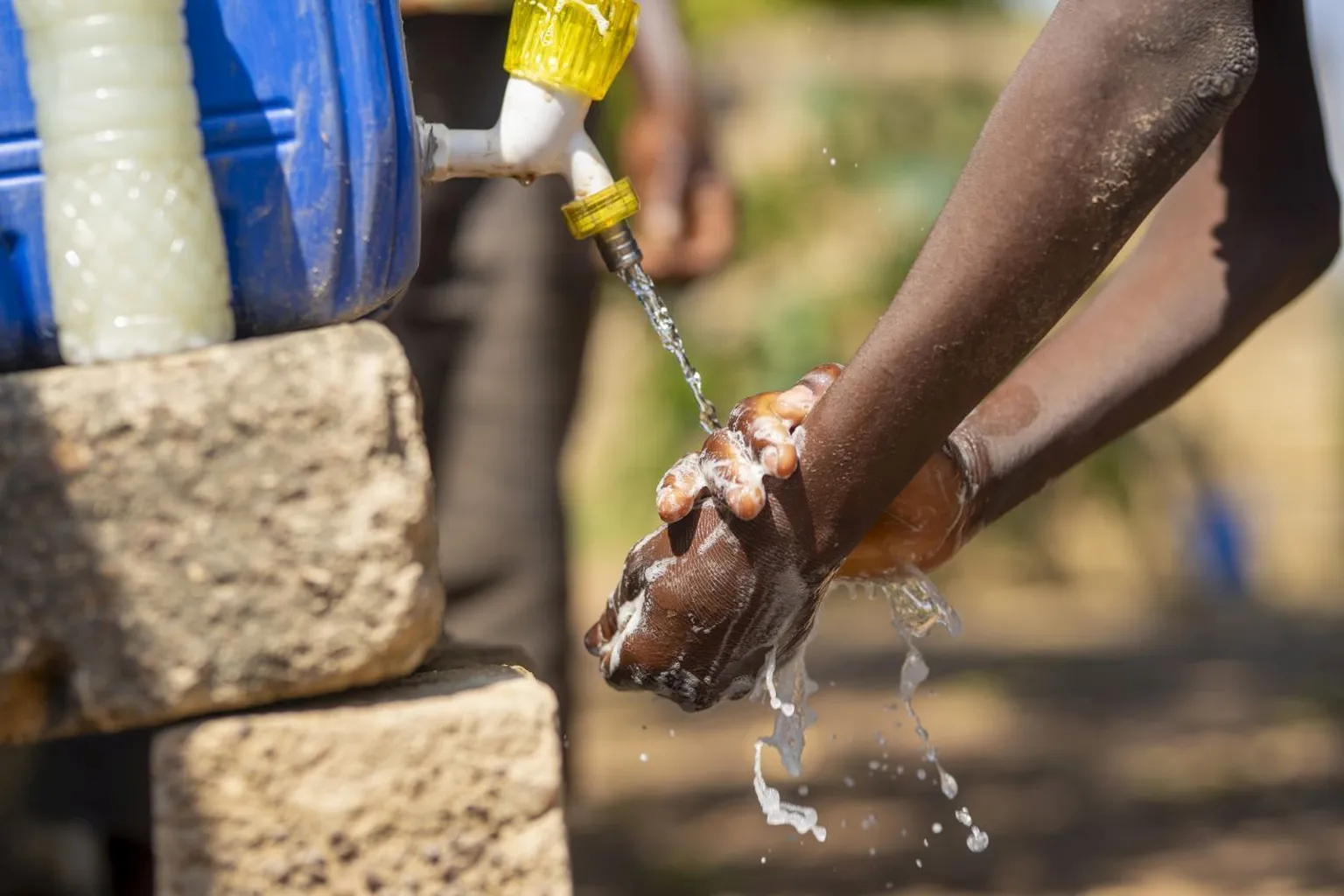 Hands being washed under a stream of water from a yellow tap attached to a blue container, with foam and bubbles visible.