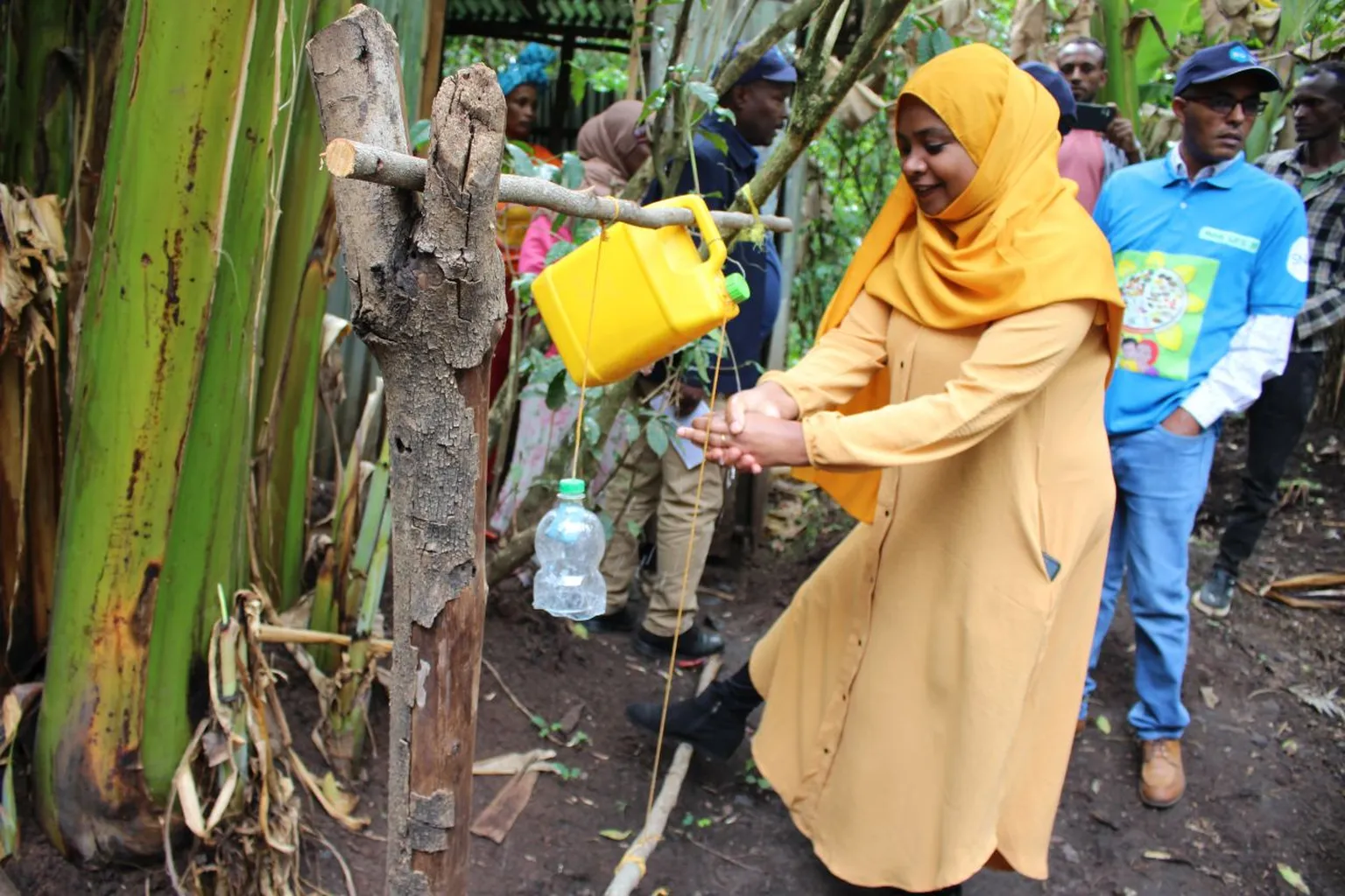 A woman in a yellow headscarf and dress uses a makeshift handwashing station outdoors, surrounded by people and greenery.