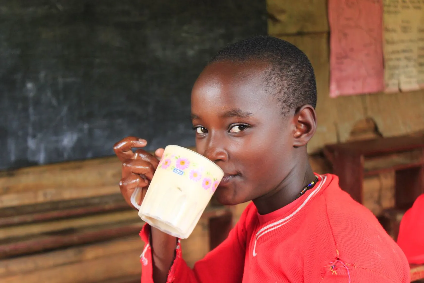 Child in red sweater sipping from a floral mug in a classroom, with a chalkboard and papers in the background.