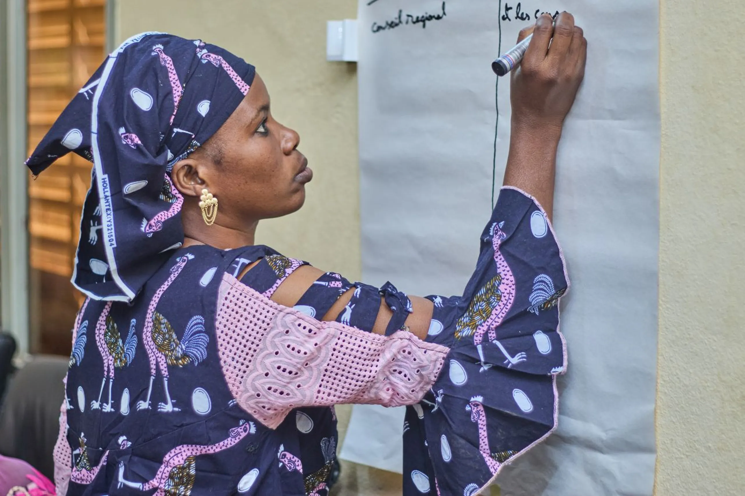 A woman in patterned attire writes on a large sheet of paper on a wall, focused on her task.