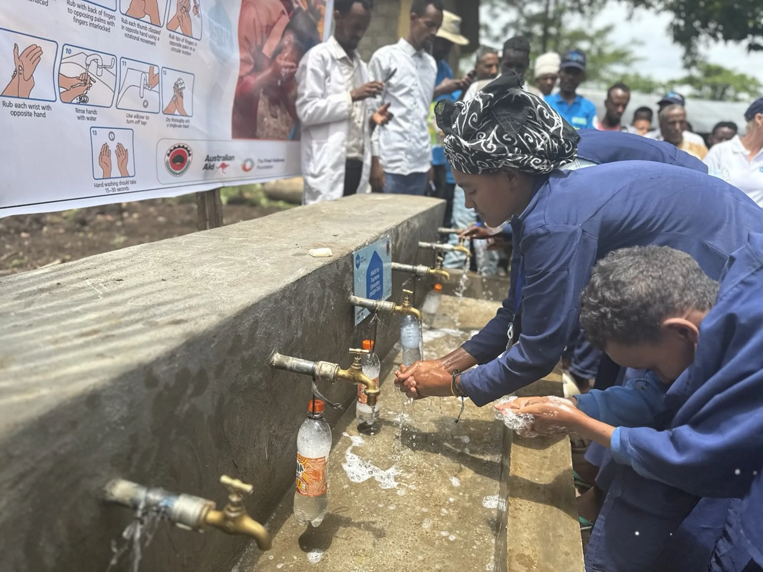 People in blue uniforms wash hands at an outdoor faucet station, with a handwashing poster and onlookers in the background.