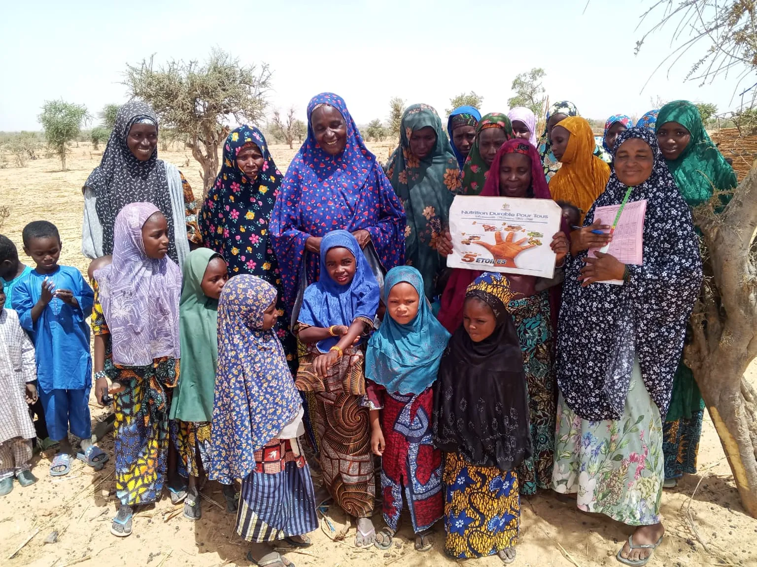 A group of women and children in colorful attire stand outdoors, some holding a sign about nutrition.