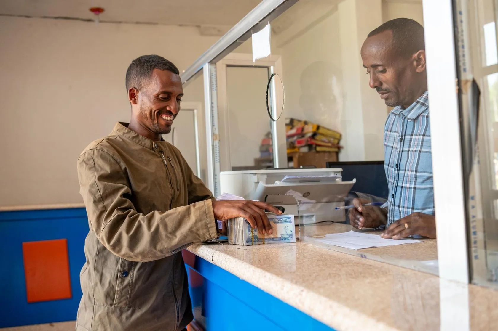 A smiling man receives money through a bank counter from a teller. The teller is seated behind protective glass in a modest office setting.