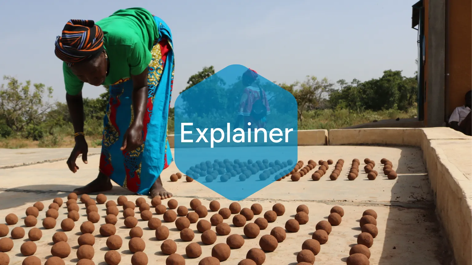 A woman in colorful clothing arranges rows of round clay balls to dry outdoors under the sun, with trees visible in the background. The word “Explainer” is overlaid in a blue hexagon at the center.