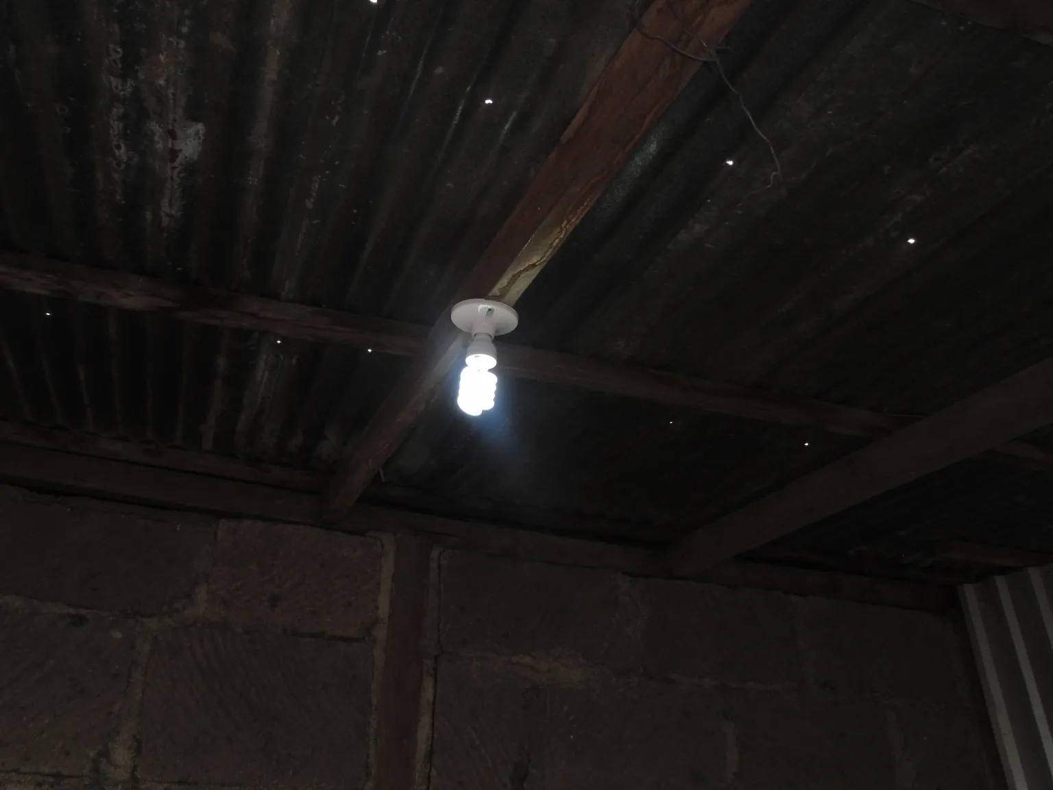 A dimly lit room with exposed wooden beams and a corrugated metal ceiling, featuring a single hanging light bulb.
