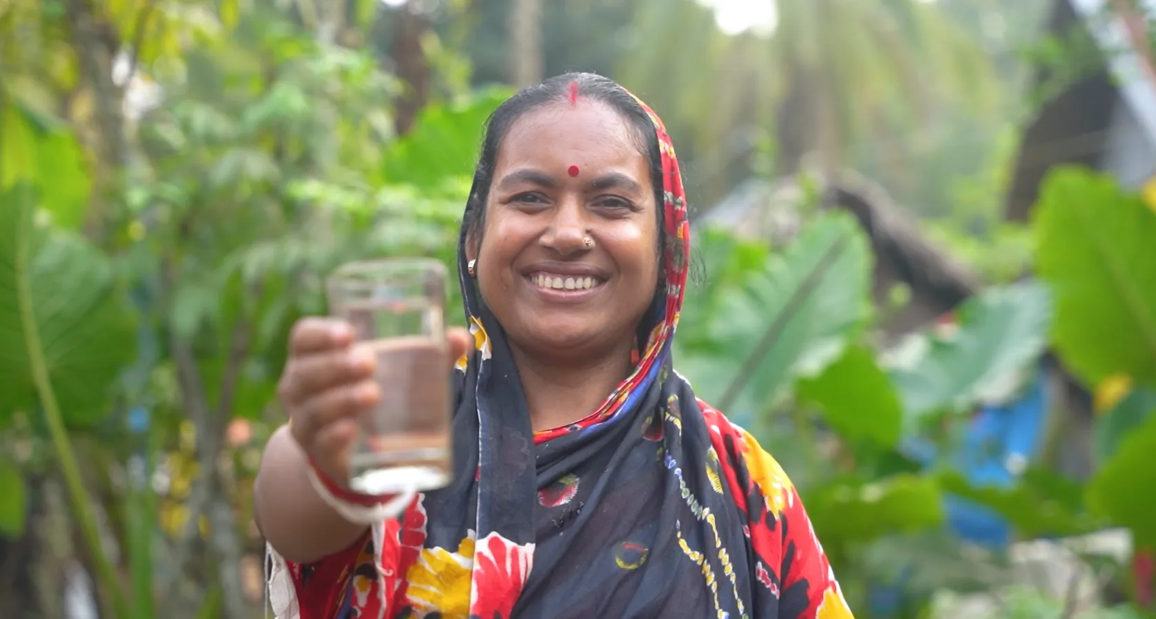 Woman in vibrant floral headscarf smiles and holds a glass of water outdoors, surrounded by lush greenery.
