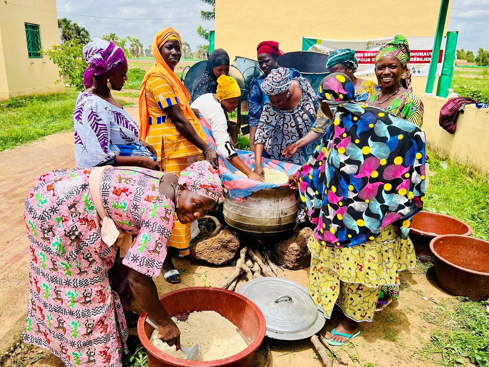 A group of women in colorful clothing are gathered outdoors, preparing a meal together with large pots and bowls.