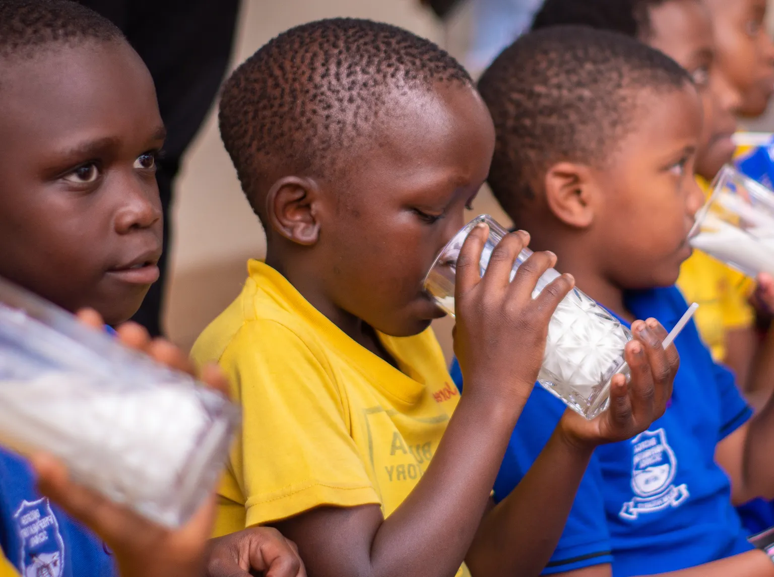 Children in colorful uniforms in a school in Uganda, sit in a row, drinking milk from glasses part of SNV's TIDE project.