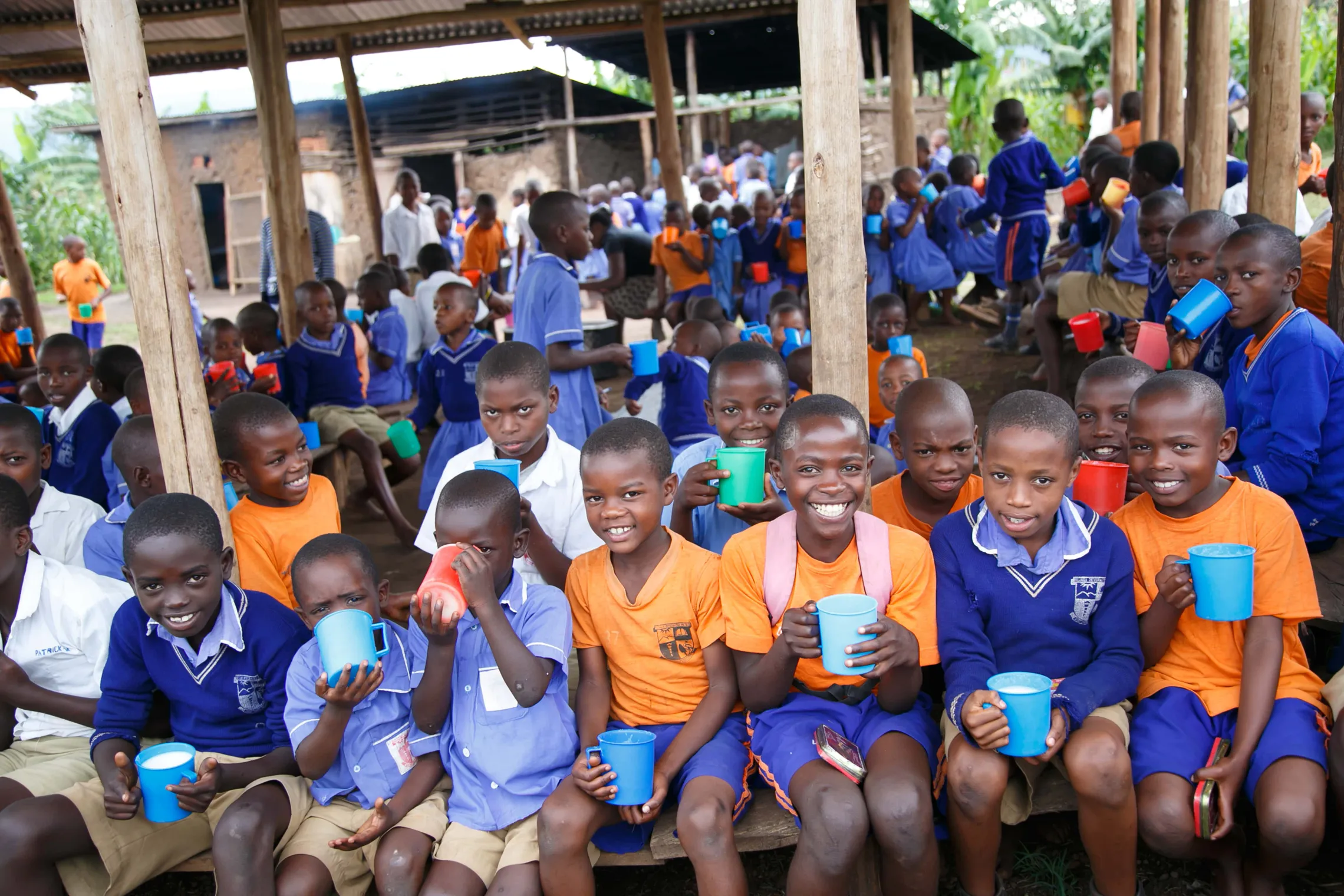 Children in colorful uniforms sit closely together under a shelter, smiling and holding brightly colored cups, creating a lively and joyful atmosphere.