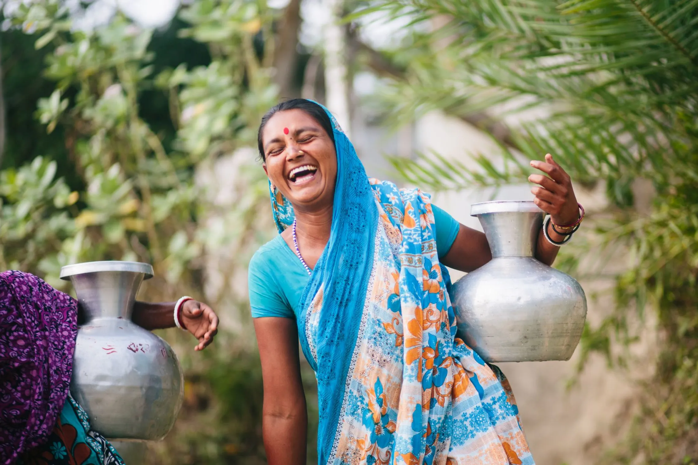 A woman in a colorful sari joyfully carrying metal water pots outdoors, surrounded by greenery.