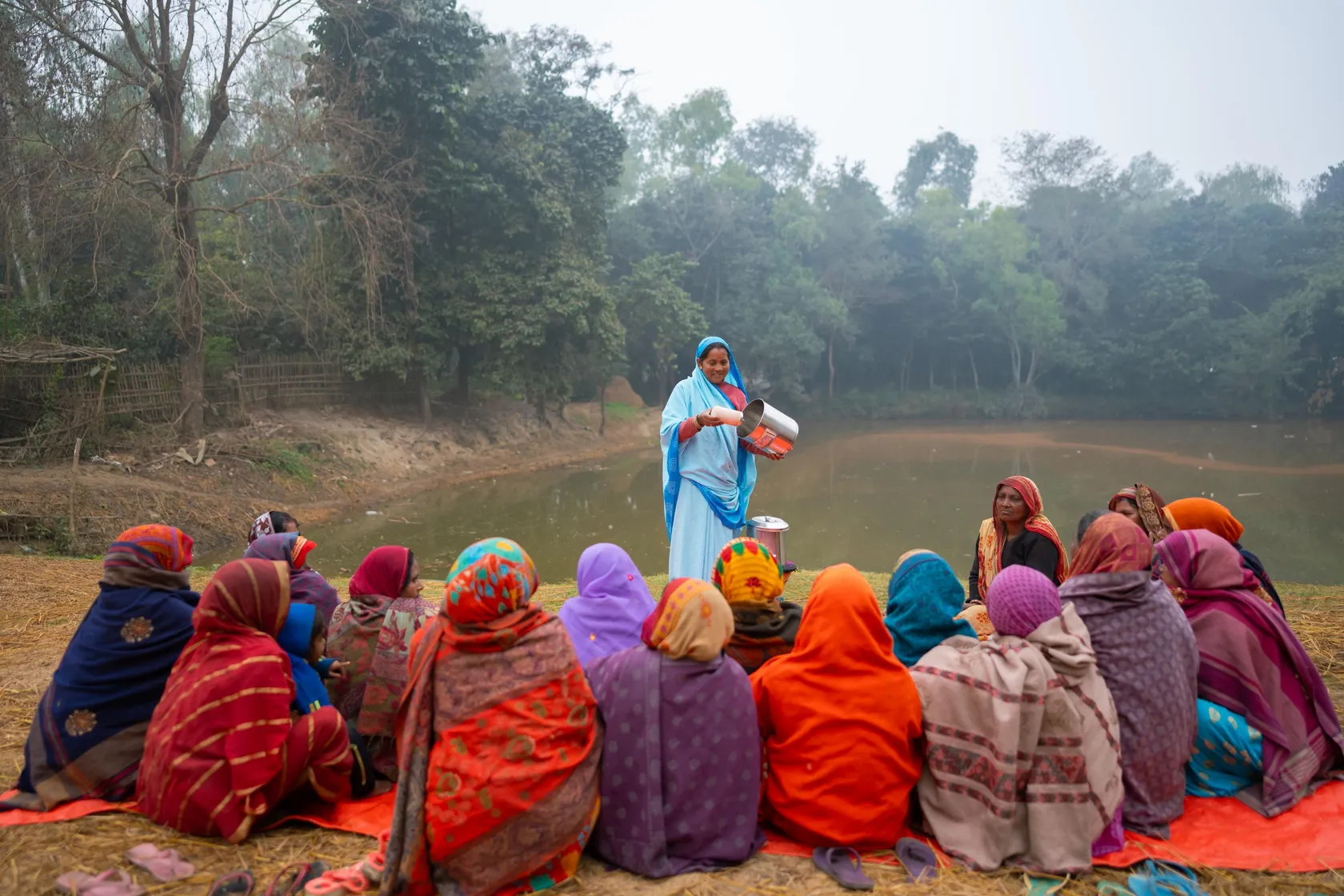 A woman in blue addresses a group seated on blankets by a pond, surrounded by trees. They wear colorful clothing and appear engaged.