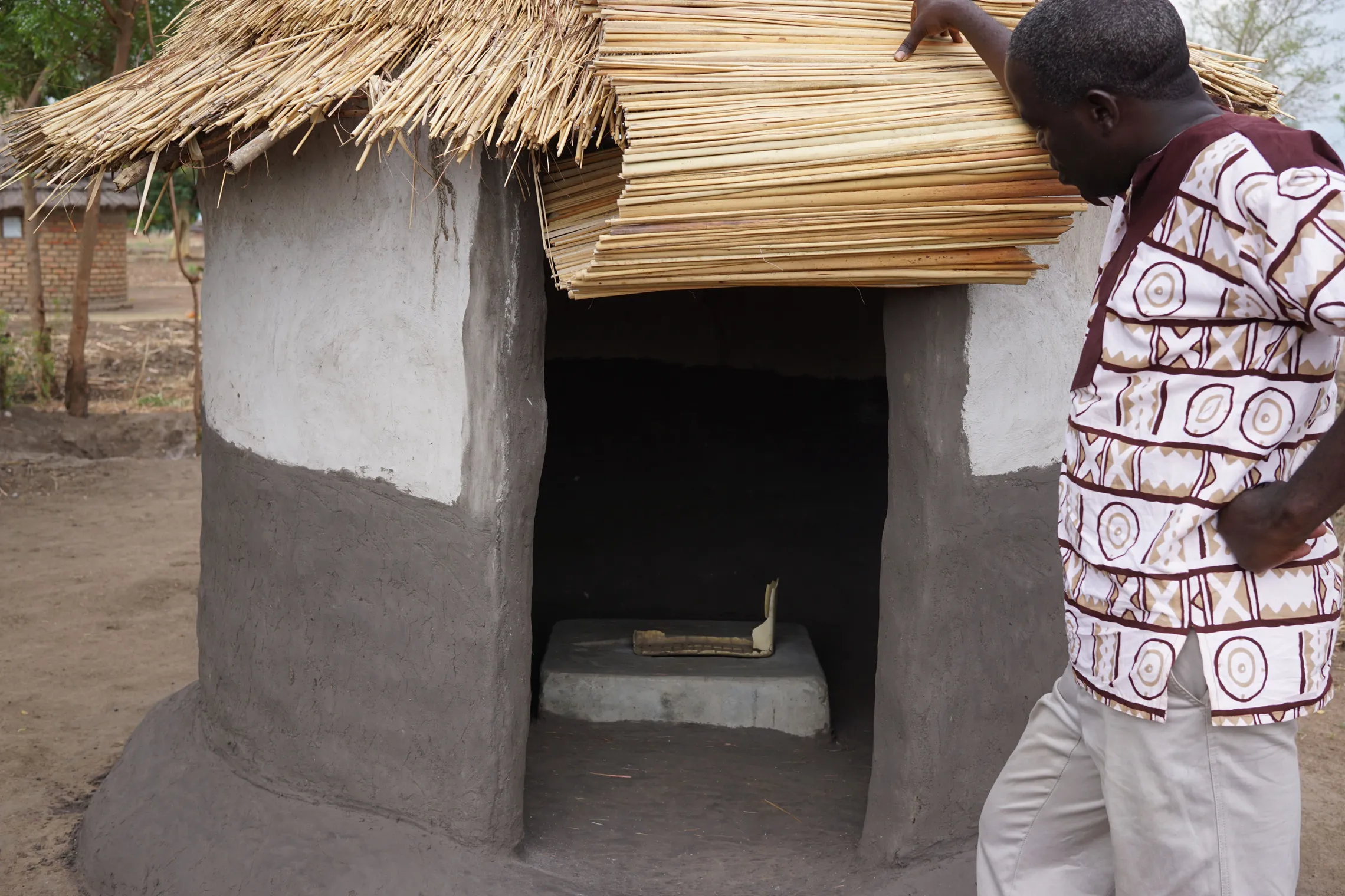 Man examines a traditional round hut with a thatched roof and mud walls, featuring a small concrete stove inside.