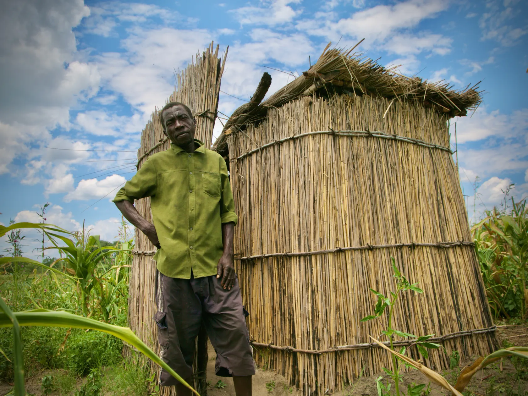 Man in green shirt stands next to two thatched huts in a field under a partly cloudy sky.