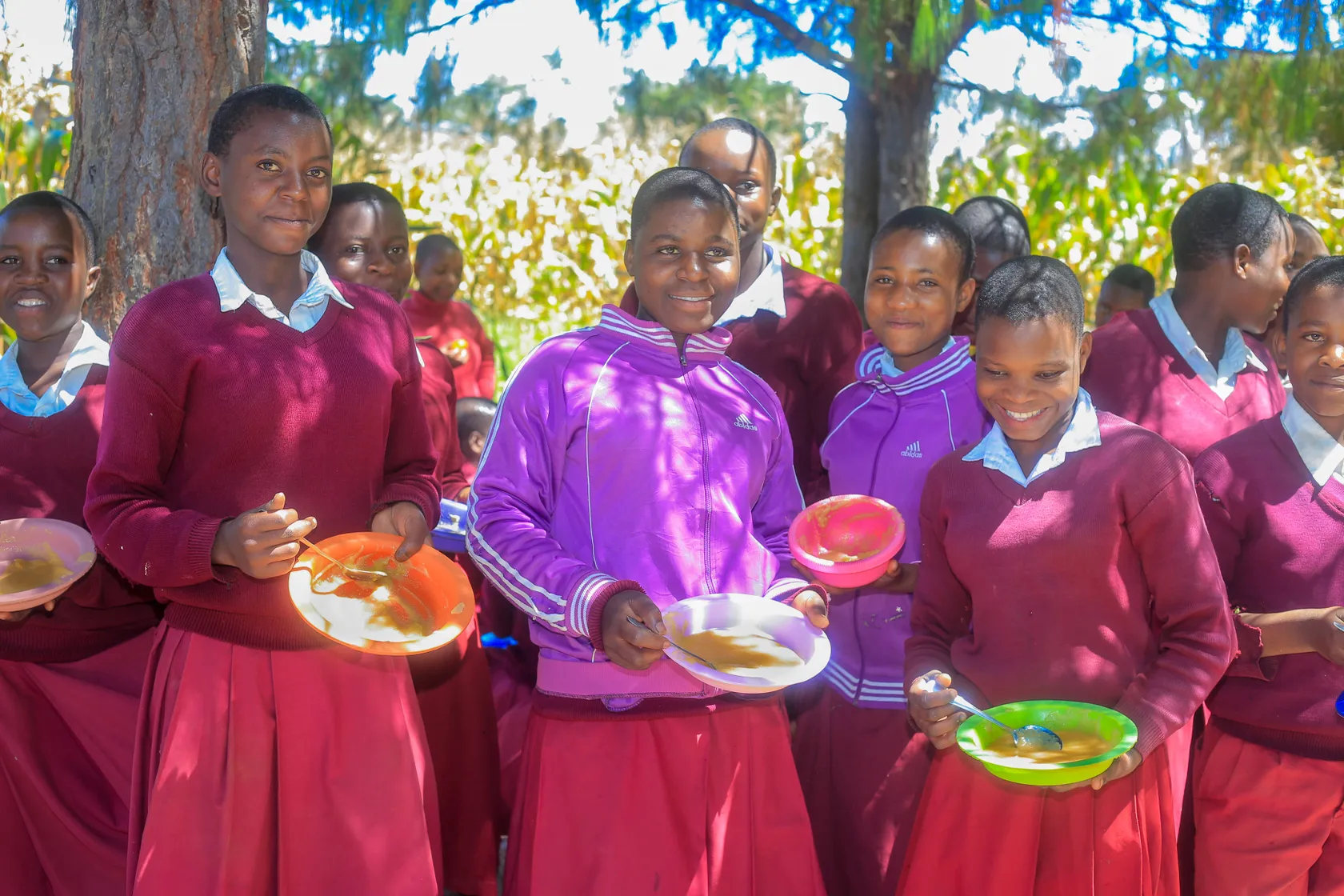 A group of students in maroon uniforms enjoy a meal outdoors, holding colorful bowls and plates, with trees in the background.