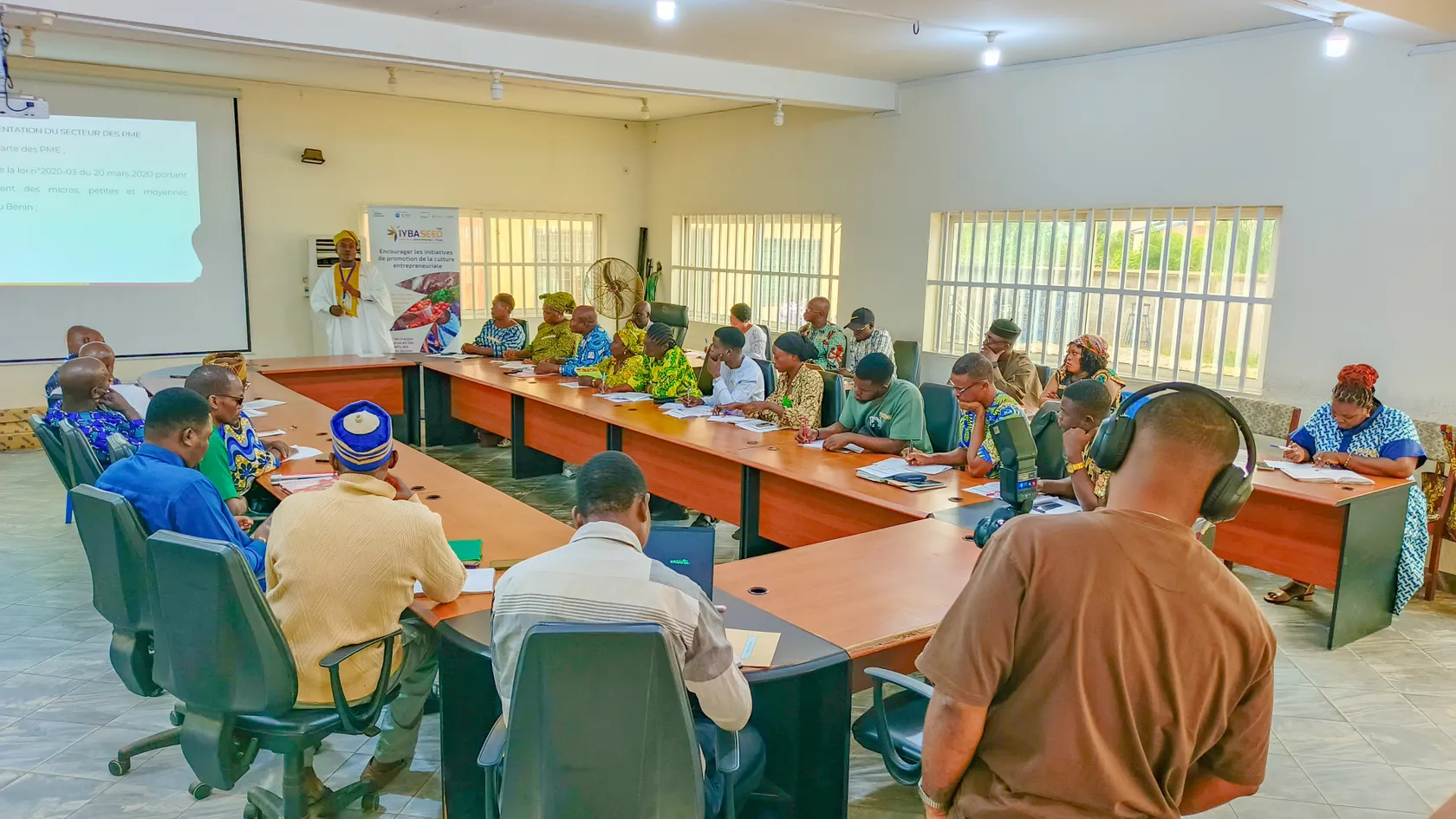 A group of people engage in a meeting around a U-shaped table in a bright room, with a presentation displayed on a screen.