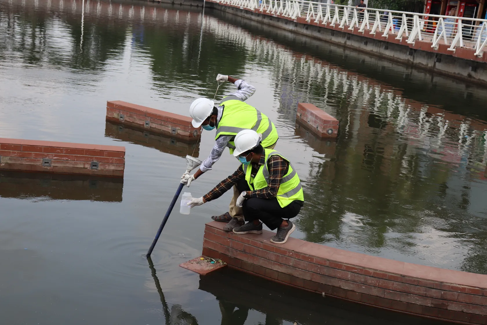 Two people in safety vests and helmets collect water samples from a pond using a pole and container, standing on brick edges.