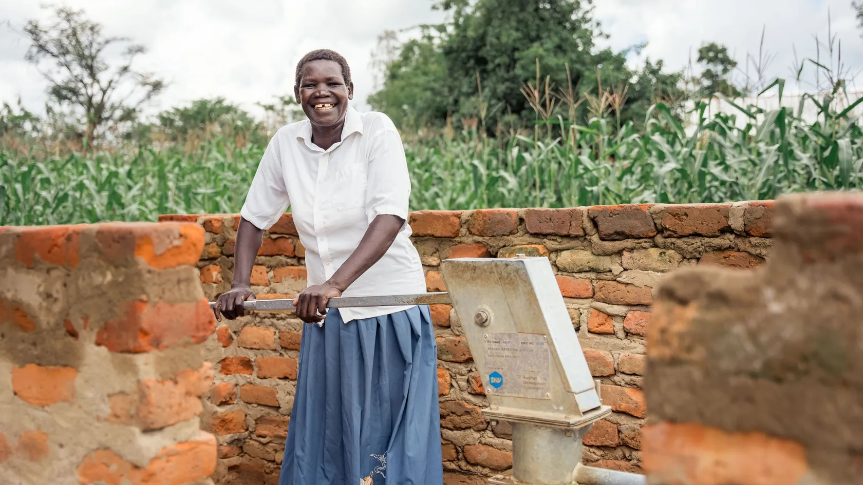 Woman smiling while operating a hand pump by a brick wall in a rural setting with greenery in the background.