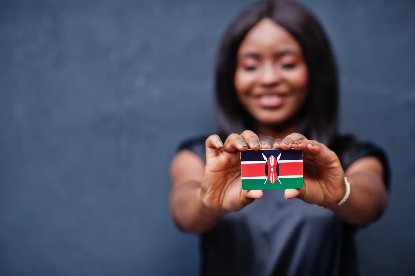 Smiling woman holding a miniature Kenyan flag card in focus, with a blurred dark background.