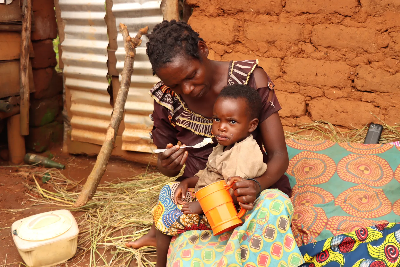 A woman feeds a young child with a spoon while the child holds an orange cup, sitting on a colorful fabric against an earthen wall.