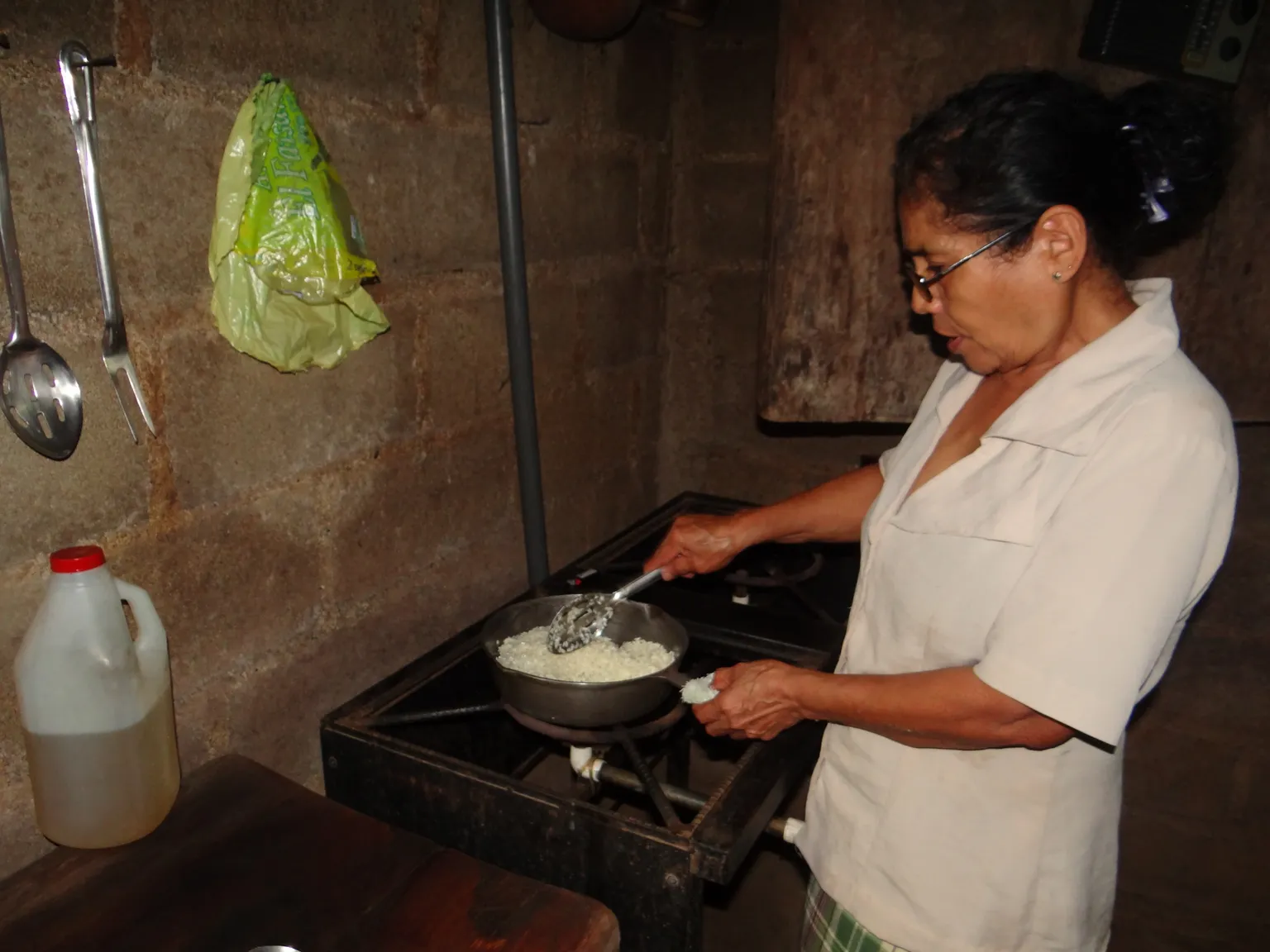 A woman in glasses cooks rice in a pan over a stove in a rustic kitchen, with utensils hanging on the wall and a plastic jug nearby.