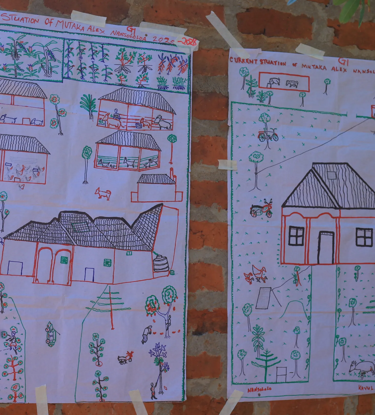 Two hand-drawn posters on a brick wall depict future and current village layouts with houses, trees, and paths, sketched in colorful markers.
