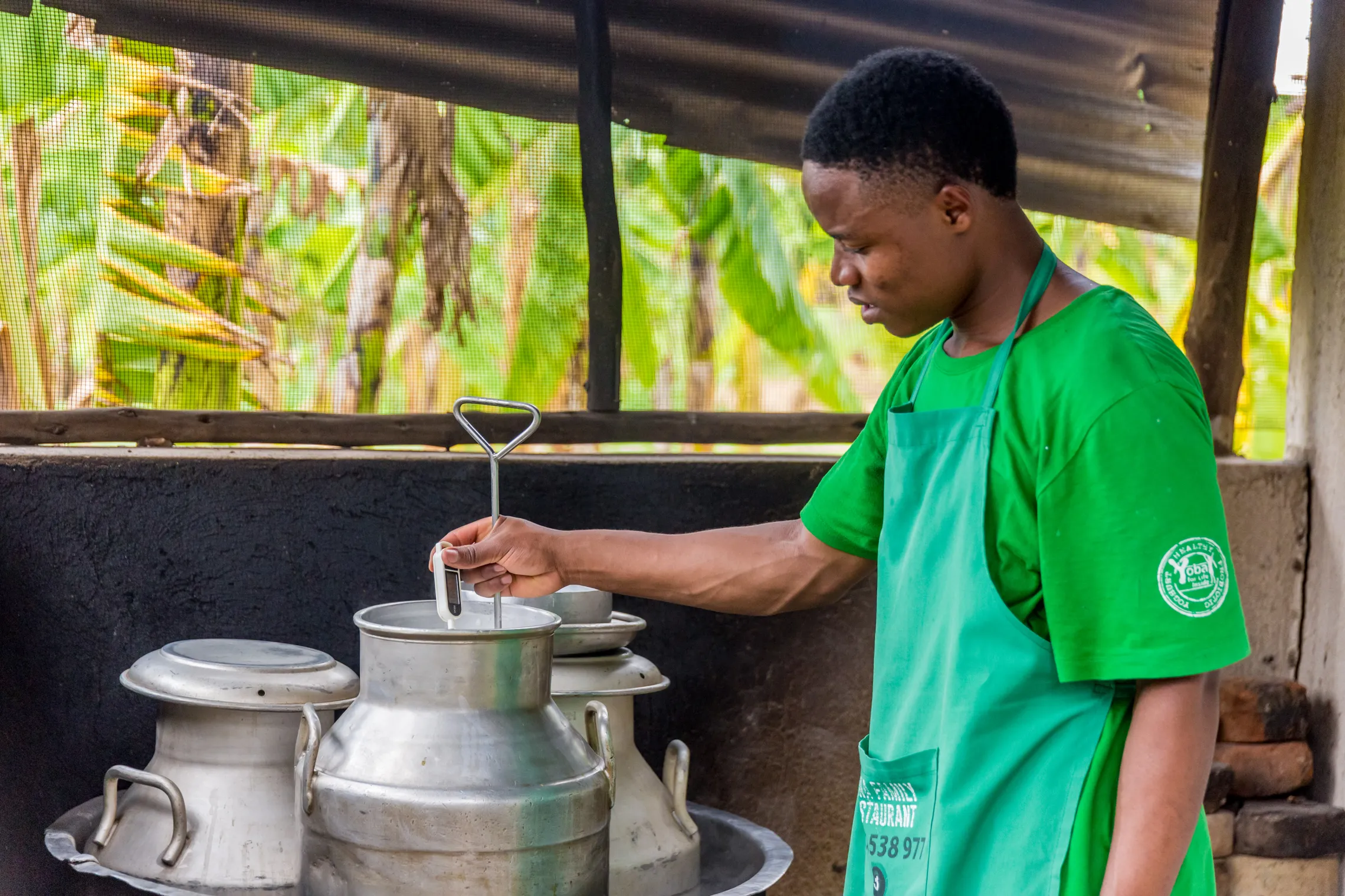 A person in a green apron stirs a pot in an outdoor kitchen to prepare school meals for students in Uganda as part of the TIDE programme.