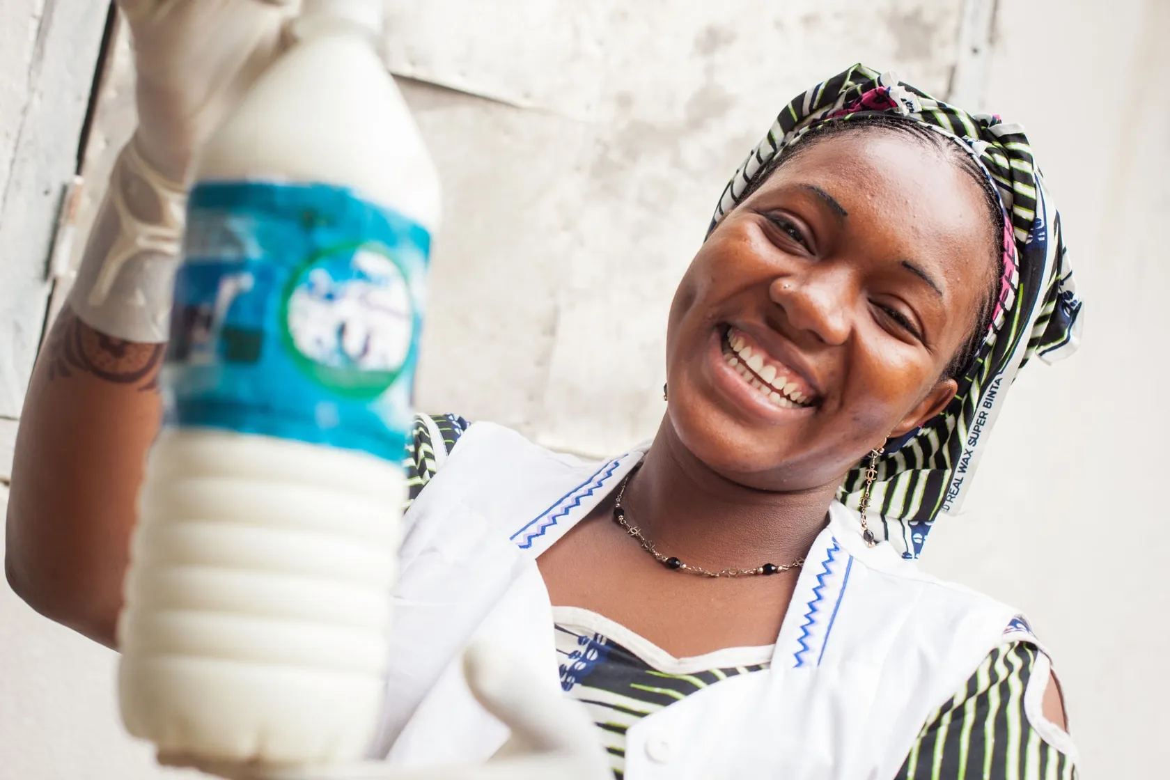 Smiling person in patterned attire and gloves holds a bottle of milk used as part of the school feeding program in East Africa