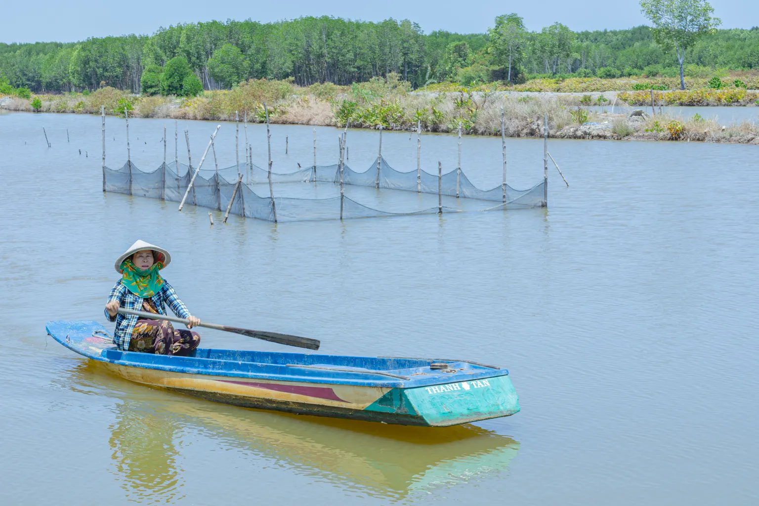 camimex vietnam - Person in a hat paddling a small boat on a calm river, with fish nets and lush greenery in the background.