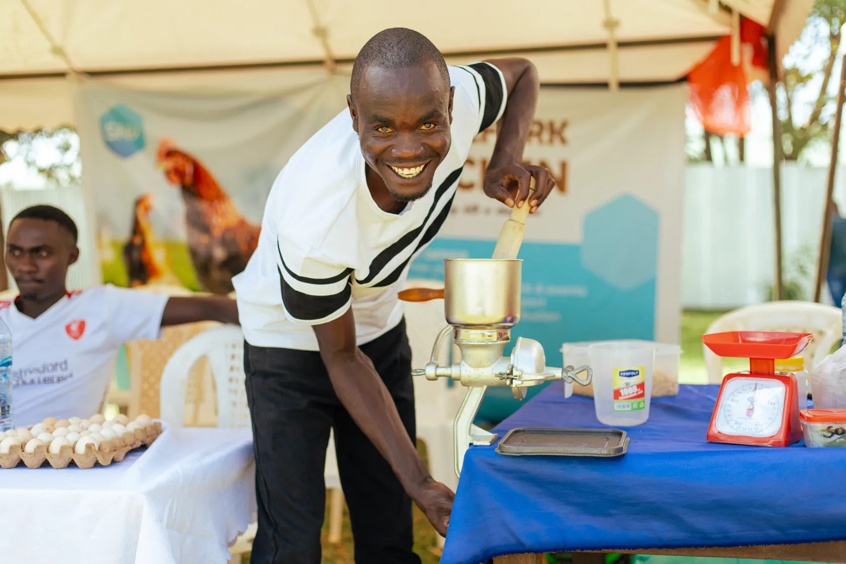 Man smiling while using a manual grinder at a market stand, with eggs, scales, and containers on the table. Another man sits nearby.