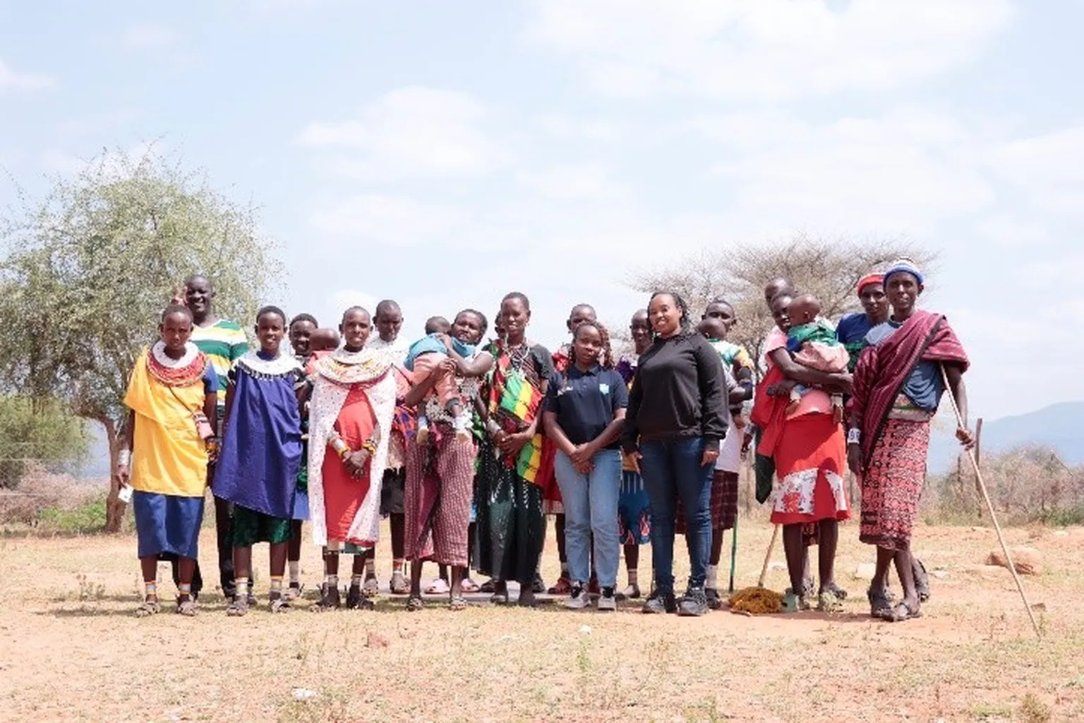 Group of adults and children standing outdoors on dry land, wearing traditional clothing and beadwork; some hold babies or walking sticks.