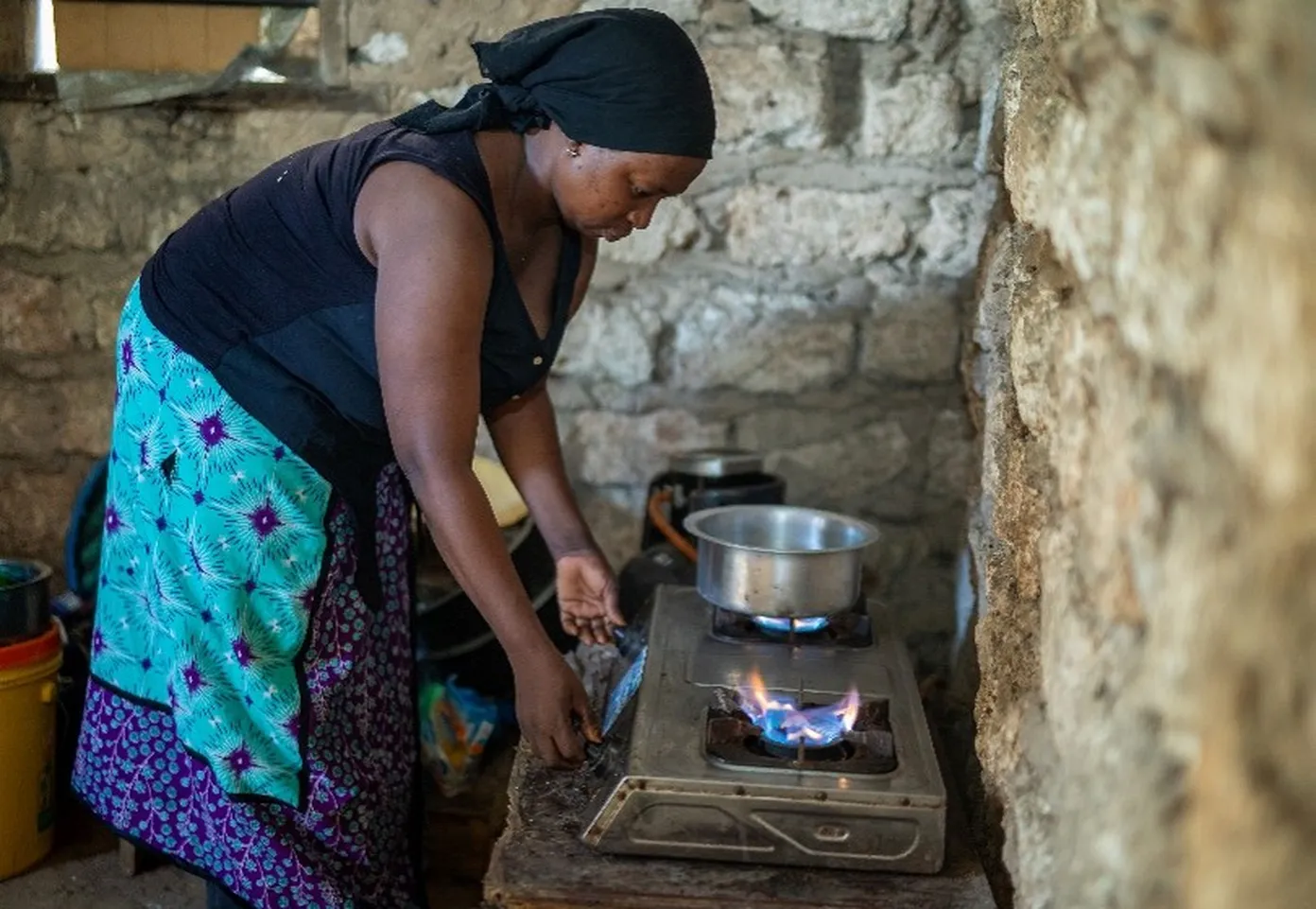 A woman in a headscarf cooks on a gas stove in a rustic kitchen, with a pot on the flame and stone walls surrounding her.