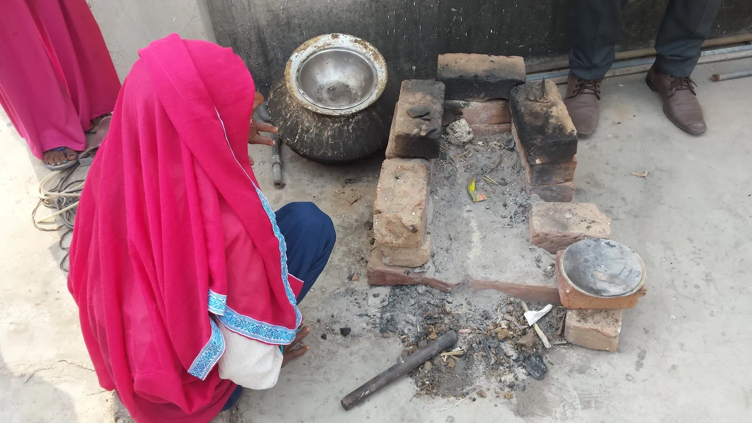 Person in a bright pink garment crouching near a makeshift outdoor stove built from bricks, with pots nearby on the ground.