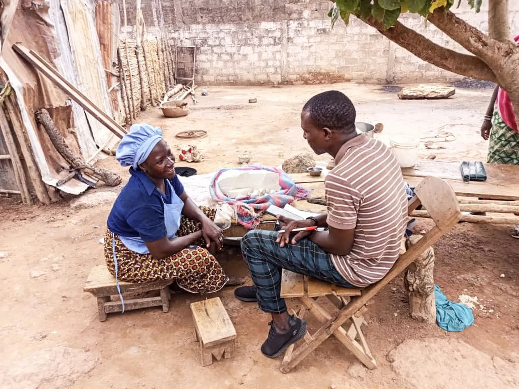 A woman in a blue headscarf talks with a man seated on wooden stools in a rustic outdoor setting with nearby tree and building materials.