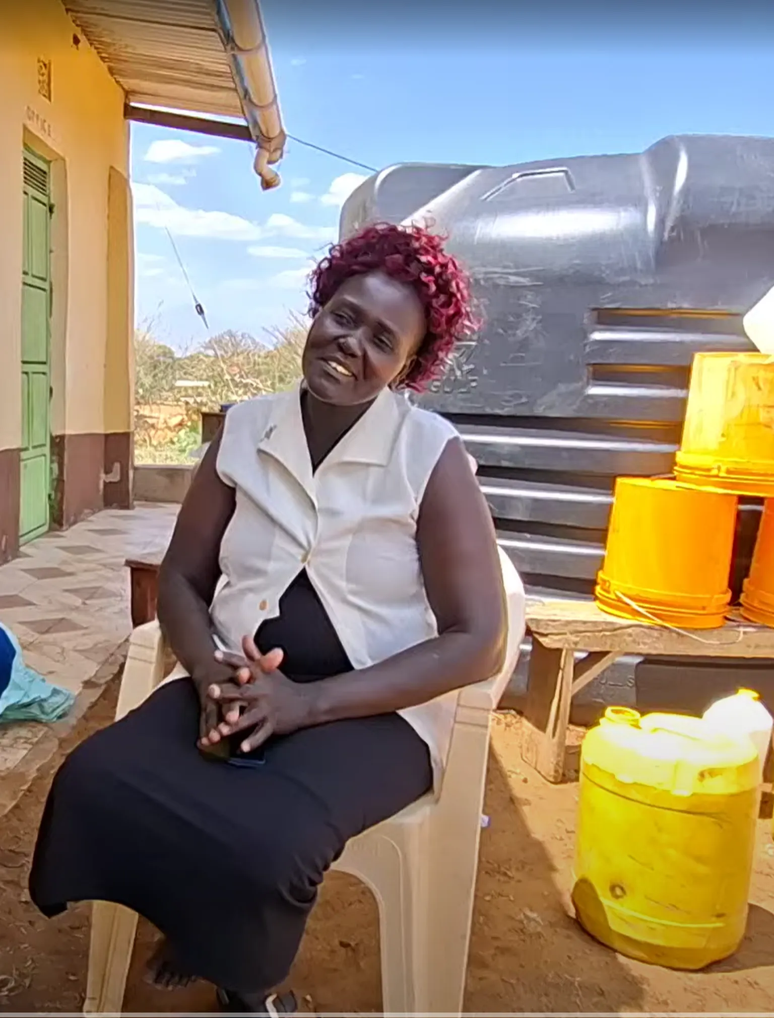 Woman sitting outdoors on a plastic chair, smiling. Behind her are a large water tank and several orange and yellow jerry cans.