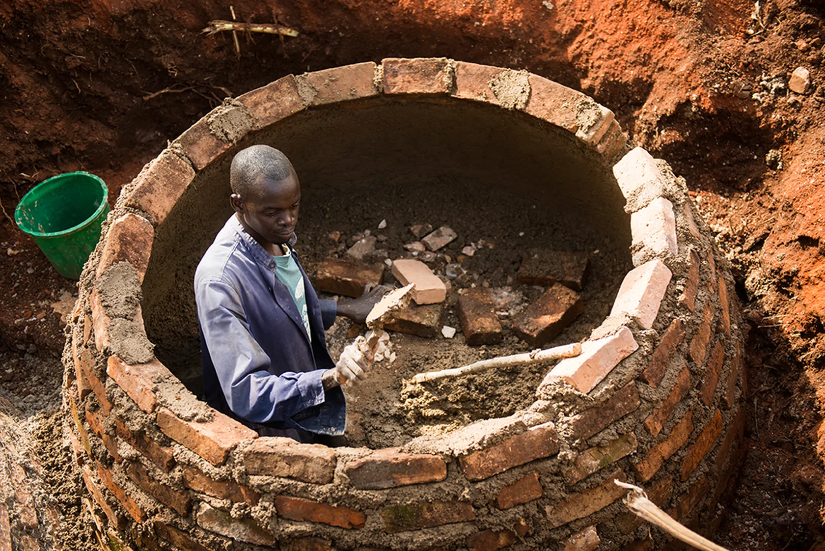 A person working inside a circular brick structure, laying bricks with mortar, surrounded by red dirt, with a green bucket nearby.