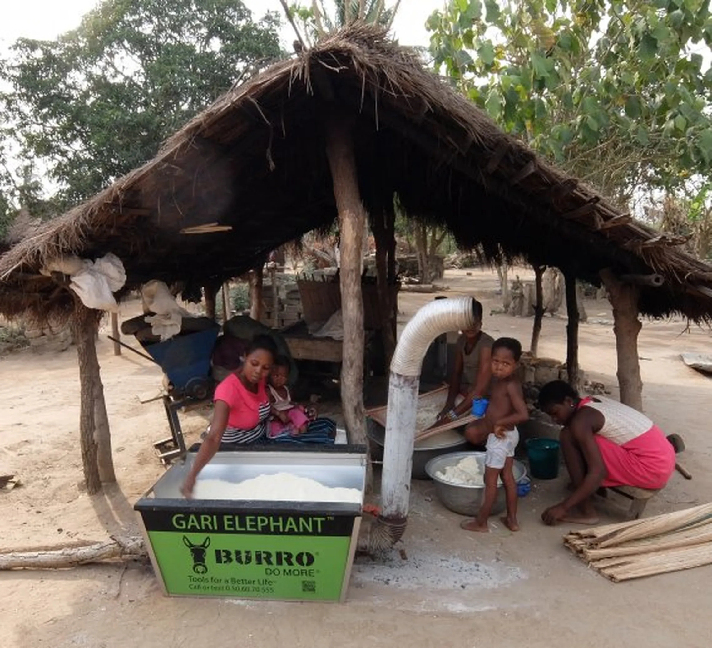 A group of people process cassava under, using a machine labeled "Gari Elephant Burro" outdoors in a rural setting in Ghana.
