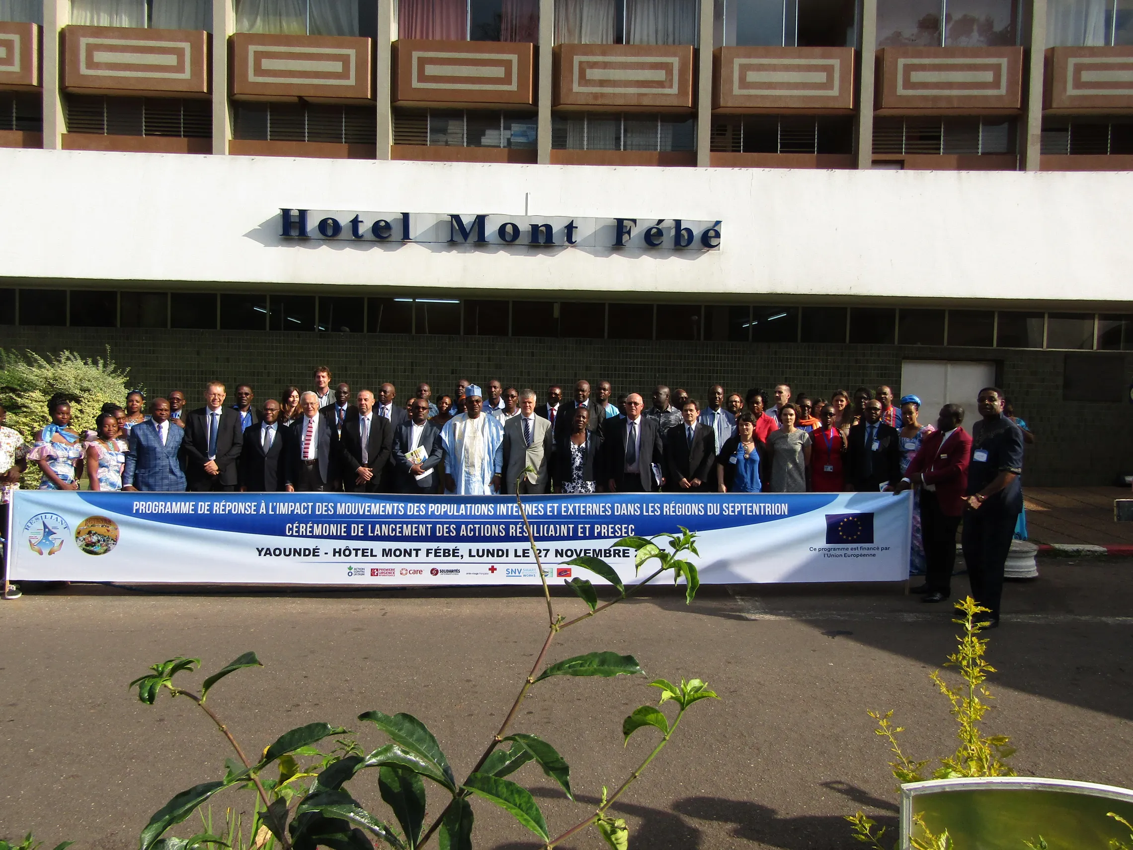 Group photo in front of Hotel Mont Fébé with attendees holding a banner for a launch event in Yaoundé, dated November 17.