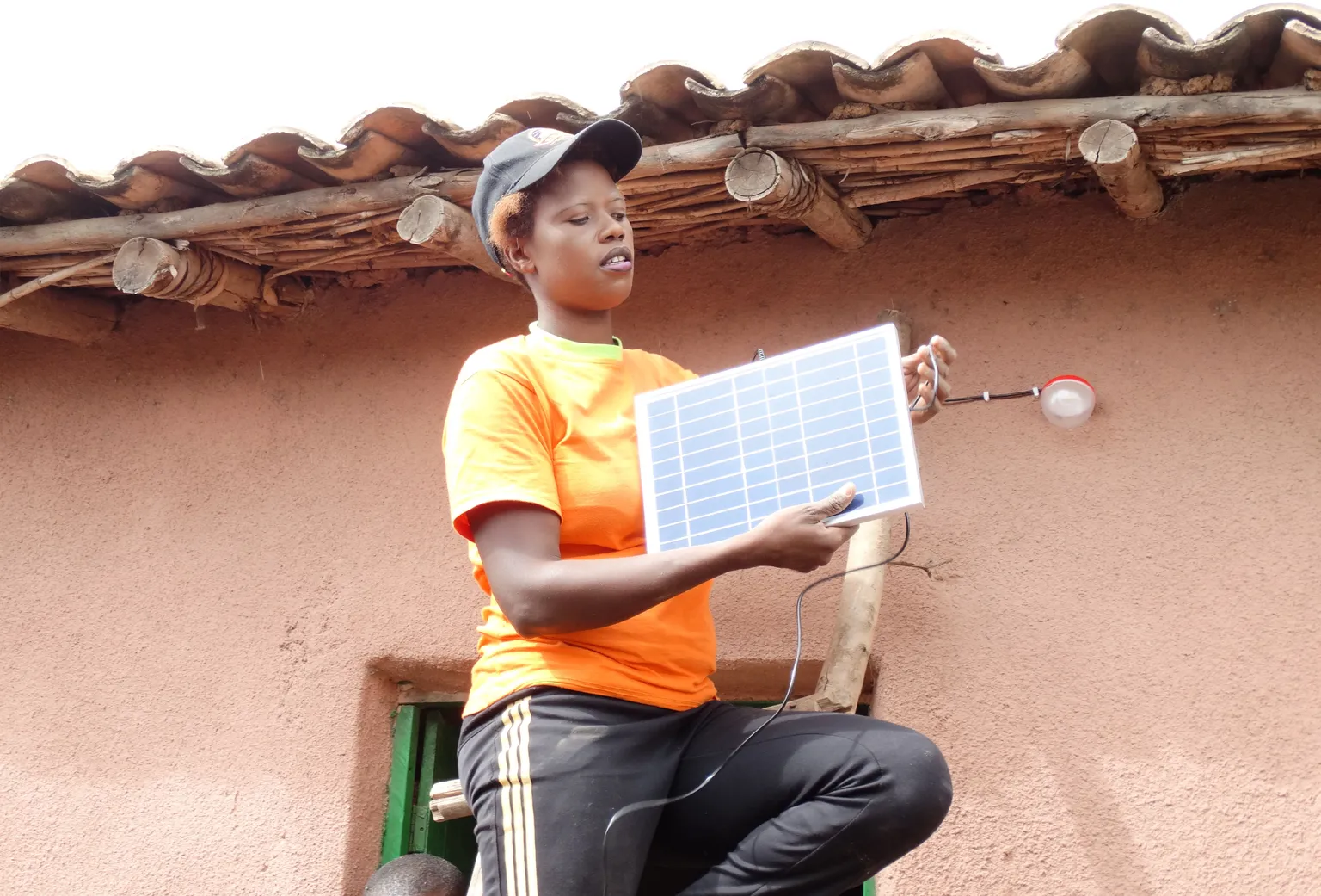 Person in an orange shirt installs a solar panel on a rustic building, holding a connected bulb. Roof made of wooden beams and tiles.