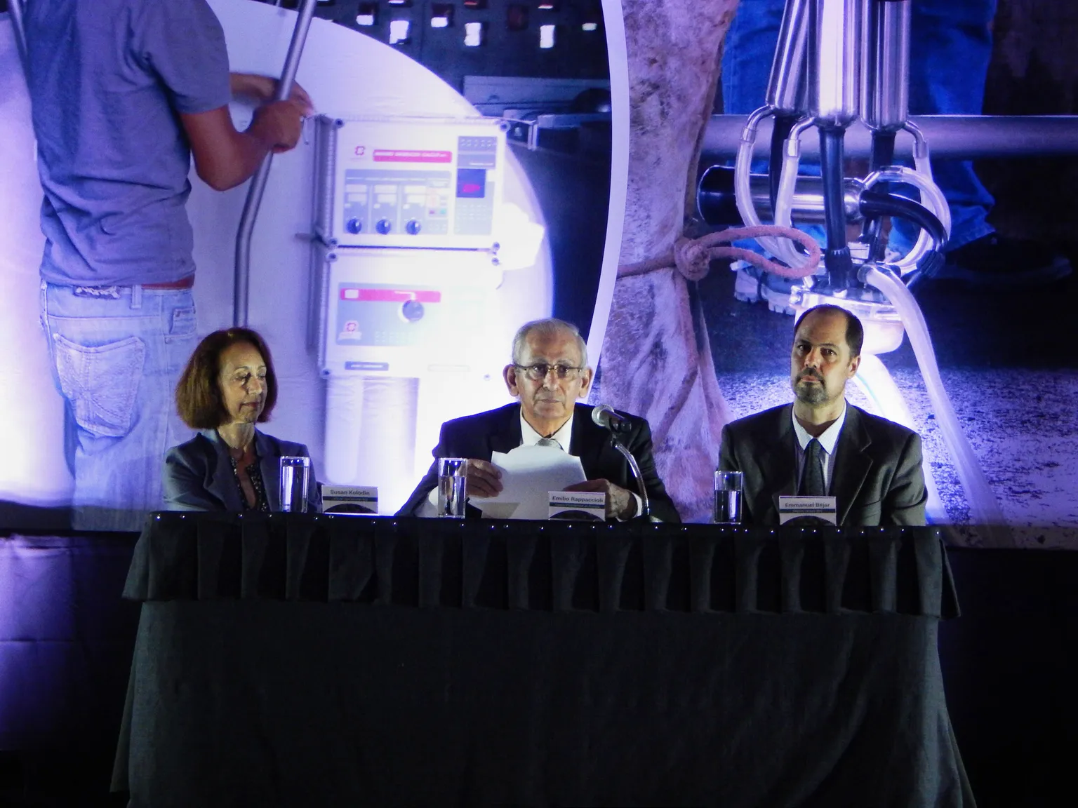 Three people sit at a conference table with microphones, against a backdrop of industrial imagery.