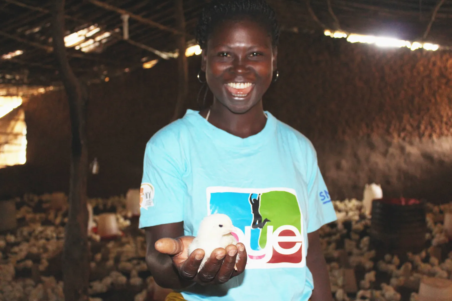 Smiling person holding a small chick in their hand, standing inside a dimly lit barn with other chicks in the background.